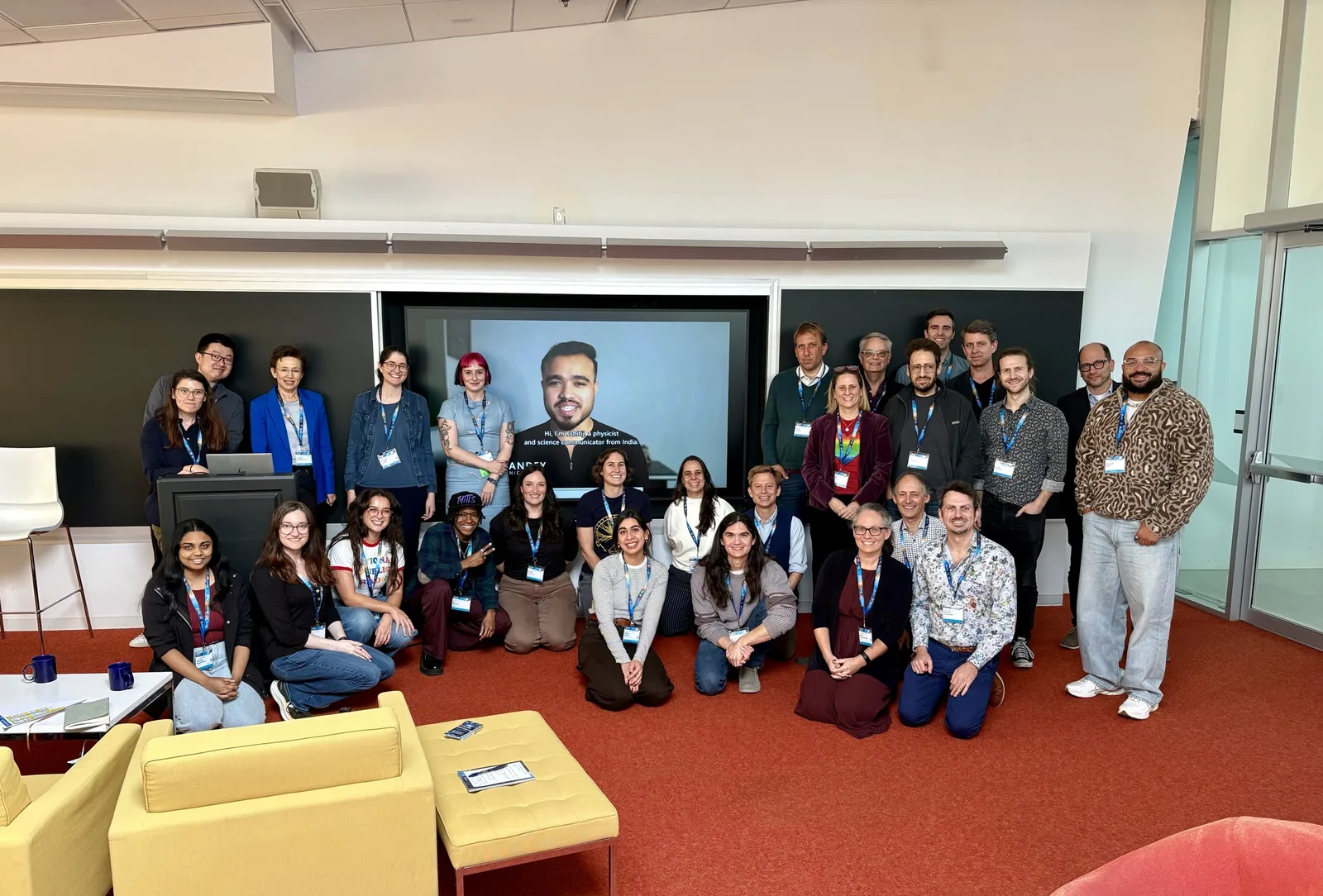 Attendees of SciComm Collider 2026 gathered in a room at the Perimeter Institute, posing in front of a large screen showing a video of Kshitij Pandey introducing himself as a physicist and science communicator from India